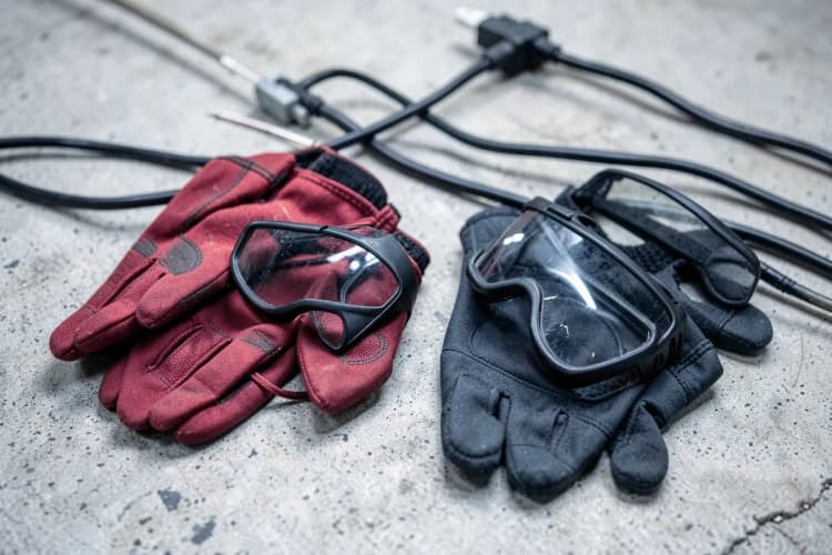 Ferramentas como luvas de proteção e óculos de segurança dispostas sobre uma superfície de concreto perto de fios elétricos.